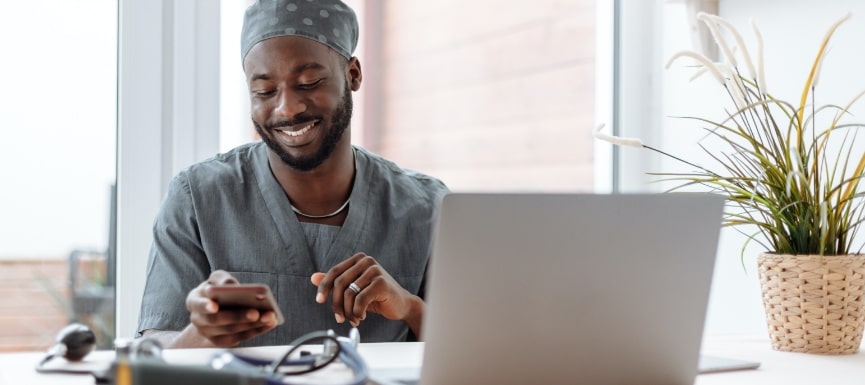 A nurse looking at their phone while sitting in front a laptop